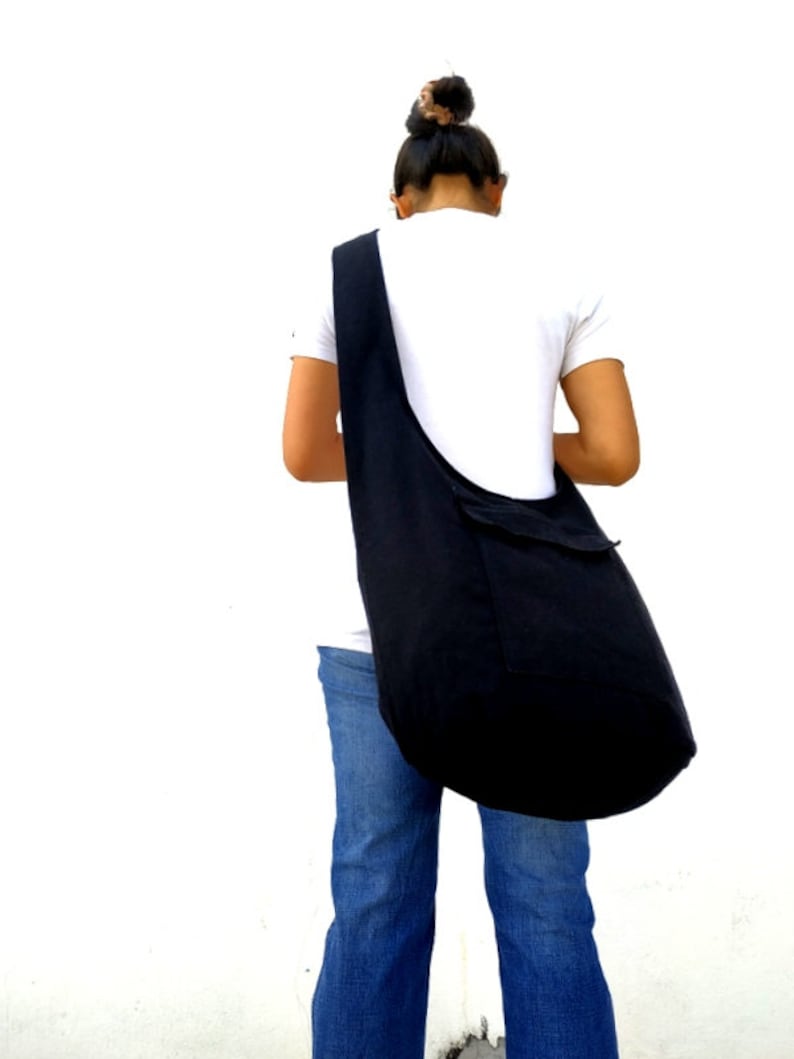 Pode incluir: Uma pessoa vestindo jeans azul e uma camiseta branca est&aacute; carregando uma grande bolsa tote de lona preta no ombro.