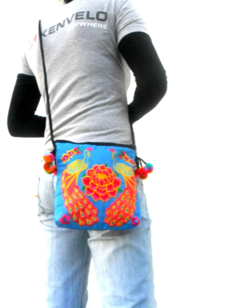 May include: A blue crossbody bag with embroidered peacocks and flowers. The bag has a long strap and colorful pom-poms.
