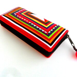 May include: A red and black zippered wallet with a colorful geometric pattern made of fabric strips. The wallet has a pink pom-pom zipper pull.
