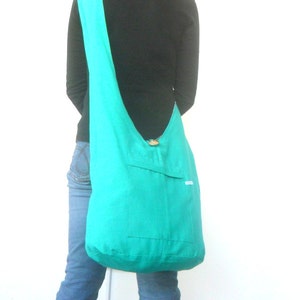 May include: A turquoise blue fabric tote bag with a single pocket on the front. The bag is worn over the shoulder of a person wearing a black shirt and blue jeans.