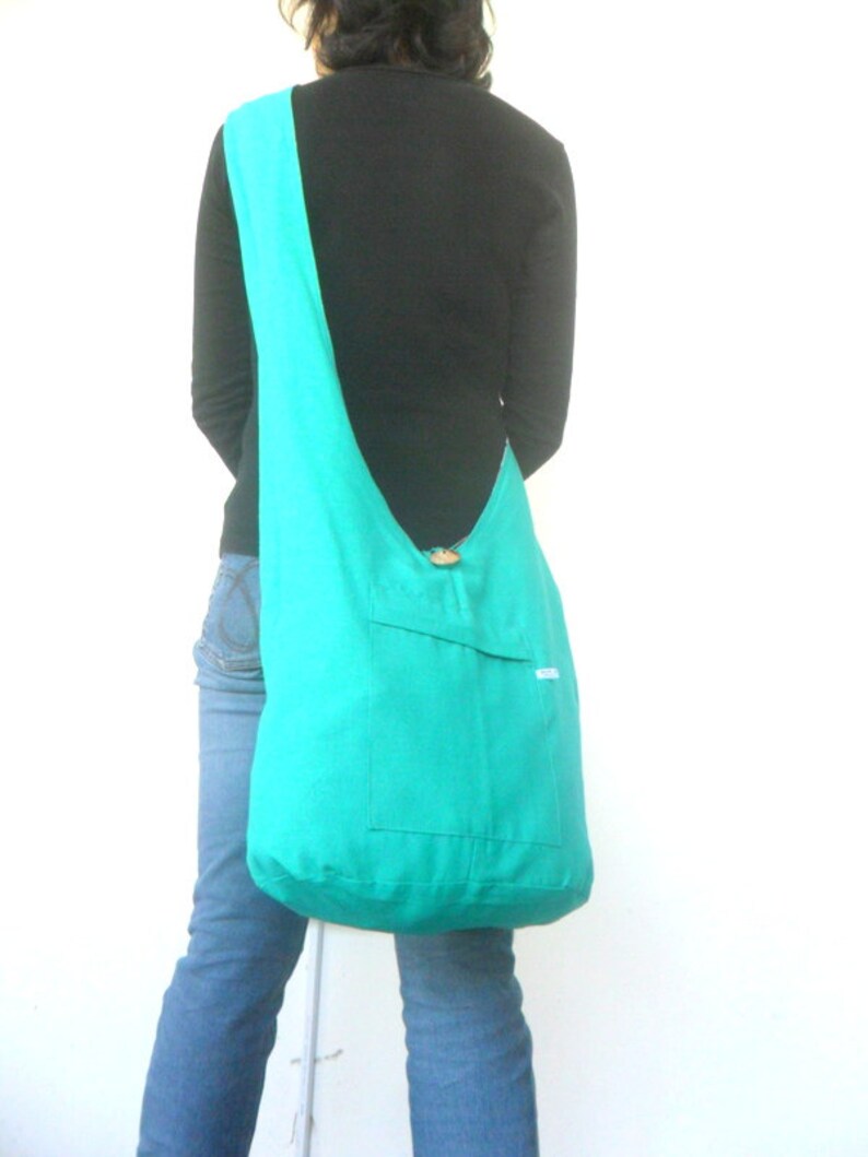 May include: A turquoise blue fabric tote bag with a single pocket on the front. The bag is worn over the shoulder of a person wearing a black shirt and blue jeans.