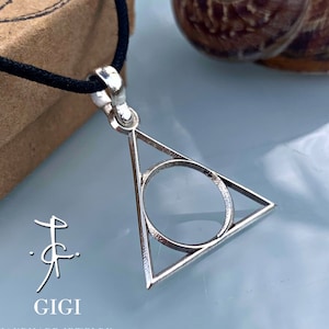 Puede incluir: Un collar con un colgante de plata con un diseño de triángulo y círculo. El colgante está en un cordón negro y está sobre una caja marrón.