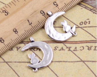 May include: Two silver-toned metal charms in the shape of a crescent moon with a cat silhouette sitting on the edge. The charms have a textured surface and a loop for attaching to a necklace or bracelet.