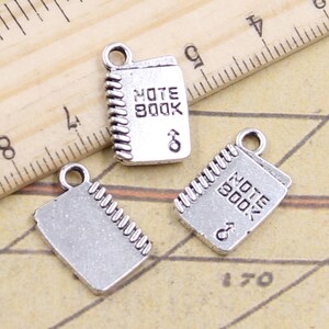 May include: Three silver charm pendants shaped like notebooks with the words "Note Book" engraved on them. The charms have a detailed design with a simulated stitching detail around the edges.
