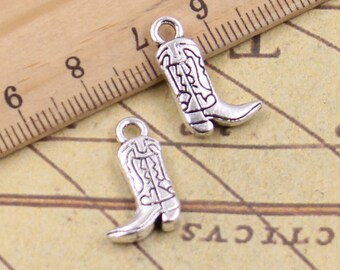 Könnte beinhalten: Zwei silberne Cowboy-Stiefel-Anhänger mit filigranen Details. Die Anhänger sind etwa 2 cm hoch und haben eine Öse oben zum Anbringen an einer Halskette oder einem Armband.