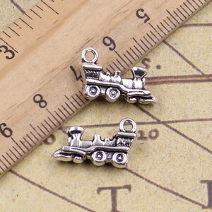 May include: Two silver-toned train engine charms with a detailed design. The charms have a loop at the top for attaching to a necklace or bracelet.
