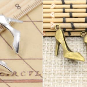 Peut inclure: Deux breloques en forme de chaussures à talons hauts, deux en argent et deux en or. Les breloques argentées sont sur une règle en bois avec des marques. Les breloques dorées sont sur une surface en bambou tissé.