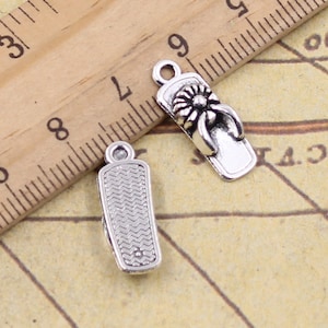 May include: Two silver charm pendants in the shape of sandals. One sandal has a flower design on the strap. The charms are on a wooden ruler with inch markings.
