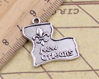 May include: A silver-toned metal charm shaped like the state of Louisiana, featuring an embossed fleur-de-lis and the text New Orleans. A wooden ruler is positioned above the charm, showing it measures approximately 3.2 centimetres wide.