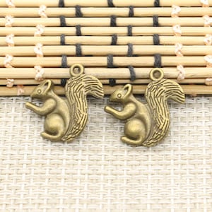 Peut inclure: Deux breloques en forme d'écureuil de couleur bronze, avec des détails de fourrure et des queues touffues. Chaque breloque a une boucle en haut pour la fixation aux bijoux ou aux travaux manuels. Les écureuils sont posés de manière ludique.