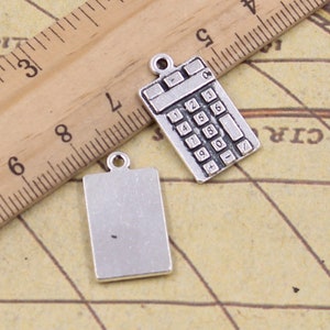 Puede incluir: Dos colgantes de plata, uno con forma de calculadora con los números del 1 al 9, 0, +, e =, y el otro un rectángulo en blanco.