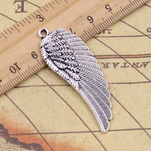 Könnte beinhalten: Silberfarbener Metallflügel-Anhänger mit detailliertem Federdesign. Der Anhänger ist etwa 6 cm lang und wird neben einem Lineal gezeigt.