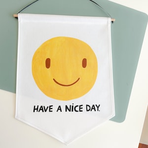 May include: White banner with a yellow smiley face and the text "HAVE A NICE DAY." The banner is rectangular with a pointed bottom and is suspended by a wooden dowel and black string.