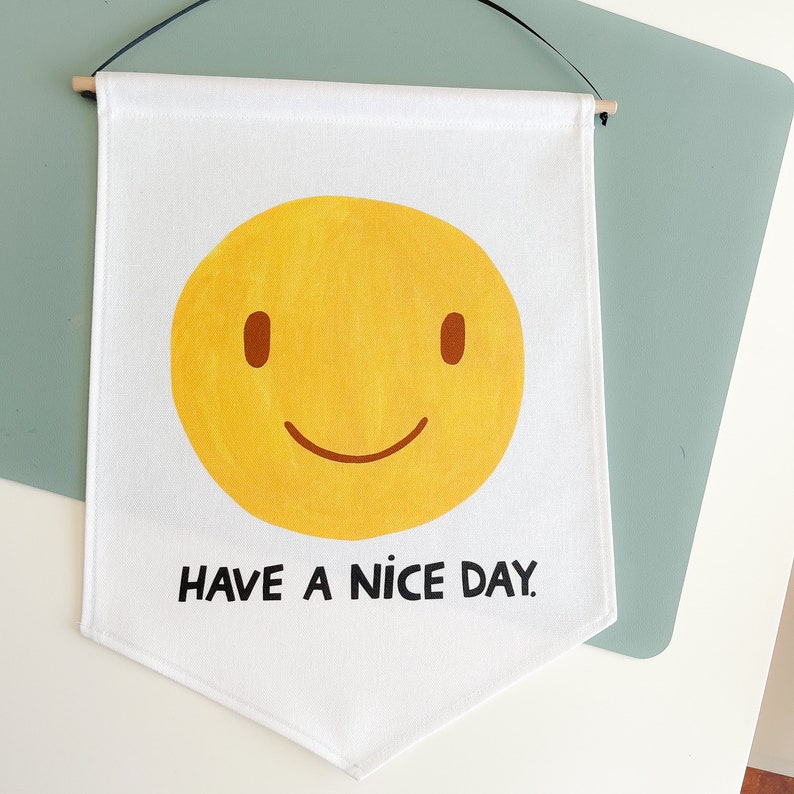 May include: White banner with a yellow smiley face and the text "HAVE A NICE DAY." The banner is rectangular with a pointed bottom and is suspended by a wooden dowel and black string.