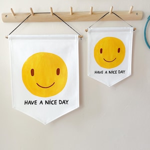 May include: Two white pennant flags with a yellow smiley face and the text "HAVE A NICE DAY." The flags are hanging from a wooden rack. The smiley face has brown eyes and a curved smile.