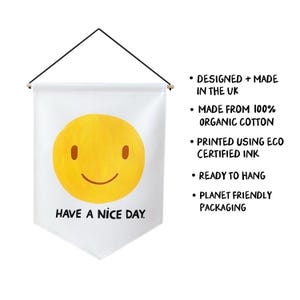 May include: A white wall hanging with a yellow smiley face and the phrase "HAVE A NICE DAY" in black text. The banner is suspended by a black cord and wooden dowels.