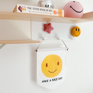 May include: A white banner with a yellow smiley face and the text "HAVE A NICE DAY" hangs below a white shelf. A pink star and a yellow smiley face are attached to the shelf. A pink smiley face lamp and a disco ball are on the shelf.