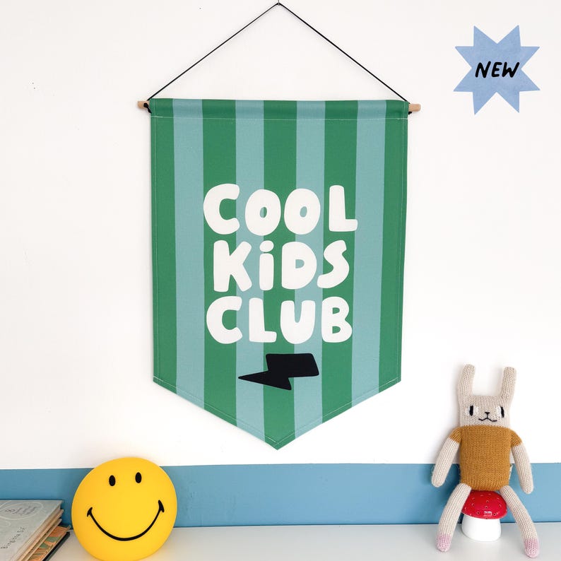 May include: Green and blue striped fabric banner with the text "COOL KIDS CLUB" in white letters and a black lightning bolt. The banner is hanging from a white wall.