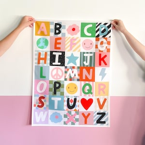 Può includere: Un colorato poster dell'alfabeto con ogni lettera in un quadrato unico. I quadrati presentano vari disegni, tra cui un globo, faccine sorridenti, stelle e simboli di pace. Il poster è tenuto contro un muro bianco e rosa.