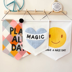 May include: Three decorative fabric banners hanging on a wooden rack. The banners feature colorful designs: a geometric pattern with the words "PLAY ALL DAY", a blue heart with the word "MAGIC", and a yellow smiley face with the words "HAVE A NICE DAY".
