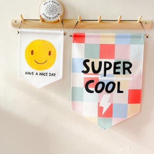 May include: Two white fabric pennants hanging on a wooden rack. The left pennant has a yellow smiley face and the text "Have a nice day." The right pennant has a colorful checkered pattern and the text "SUPER COOL" with a lightning bolt.