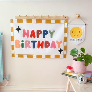 May include: A white banner with the words "HAPPY BIRTHDAY" in colorful letters, accented with black stars. The banner is framed with a yellow and white striped border and hangs on a wooden rack. A smaller banner with a smiley face and the text "HAVE A NICE DAY" is also displayed.