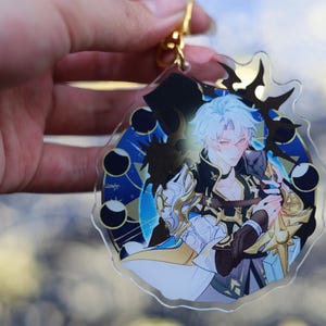 May include: Clear acrylic keychain featuring a detailed anime-style illustration of a character with white hair and a black and gold outfit. The design includes a blue and gold celestial background and a gold-colored clasp.