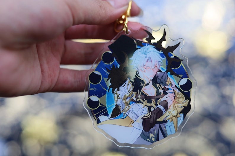 May include: Clear acrylic keychain featuring a detailed anime-style illustration of a character with white hair and a black and gold outfit. The design includes a blue and gold celestial background and a gold-colored clasp.