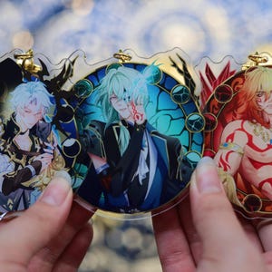 May include: Three anime-style acrylic charms featuring male characters. Each charm has a circular design with a gold-colored top loop. The characters have different hair colors and outfits, set against colorful backgrounds with decorative borders.