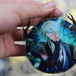May include: A clear acrylic charm featuring an anime-style character with light green hair and red eyes. The character is set against a blue and teal background with a decorative border. A gold-colored keychain is attached.