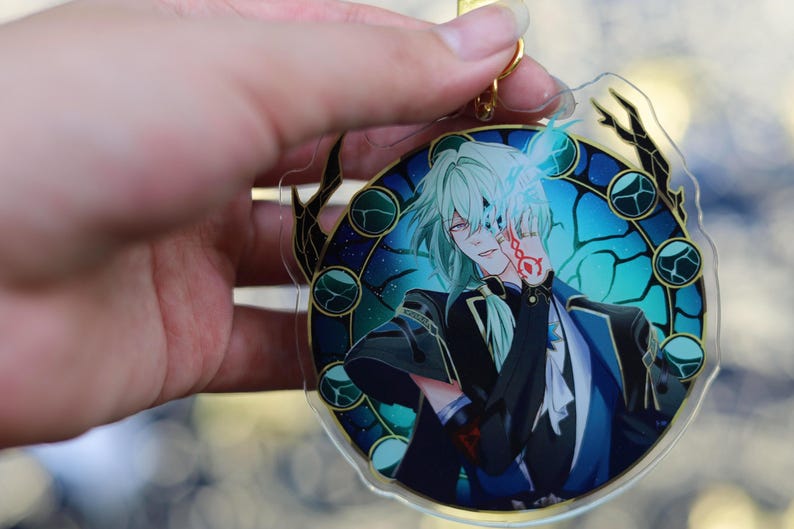 May include: A clear acrylic charm featuring an anime-style character with light green hair and red eyes. The character is set against a blue and teal background with a decorative border. A gold-colored keychain is attached.