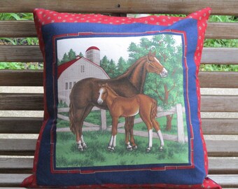 May include: Square decorative pillow featuring a pastoral scene with two horses in a field, a white barn, and a red roof. The pillow has a blue border and a red paisley patterned edge. The pillow is placed on a wooden bench.
