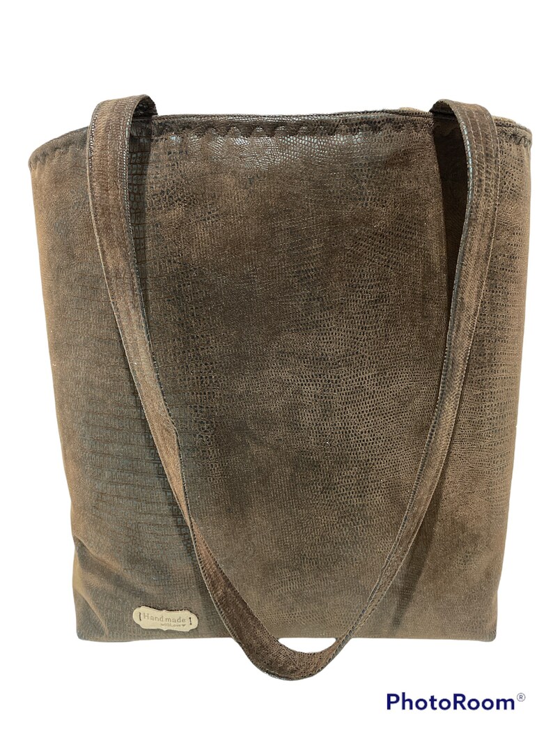 K&ouml;nnte beinhalten: Eine braune Ledertasche mit zwei Henkeln. Die Tasche hat eine strukturierte Oberfl&auml;che und ein kleines Etikett mit der Aufschrift "Handmade".