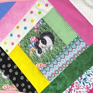 May include: A colorful patchwork quilt featuring a central panel with two black and white rabbits in a grassy field. The quilt incorporates various patterned fabrics, including polka dots, gingham, and floral designs, in shades of pink, blue, green, and yellow.