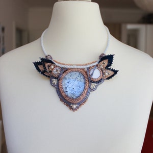 May include: A white beaded necklace with a large, oval, blue and white agate stone pendant. The pendant is surrounded by smaller white beads and black and gold beads. The necklace is on a white mannequin.