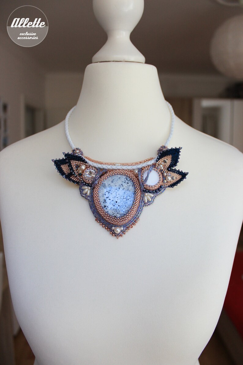 May include: A white beaded necklace with a large, oval, blue and white agate stone pendant. The pendant is surrounded by smaller white beads and black and gold beads. The necklace is on a white mannequin.