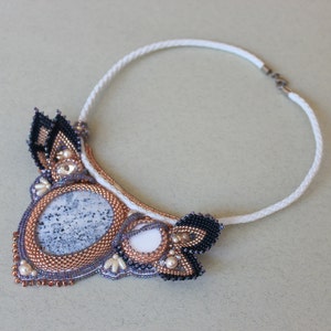 May include: A white beaded necklace with a large, gray, beaded pendant. The pendant is surrounded by smaller black, white, and gold beads. The necklace is made with a white cord and has a silver clasp.