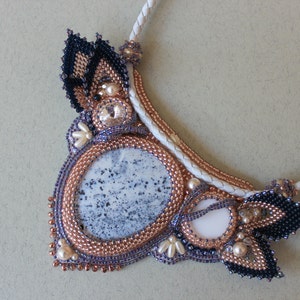 May include: A white leather cord necklace with a large, oval-shaped, gray and white agate stone pendant. The pendant is surrounded by a beaded frame with copper, gold, and black beads. The necklace is a statement piece with intricate details.