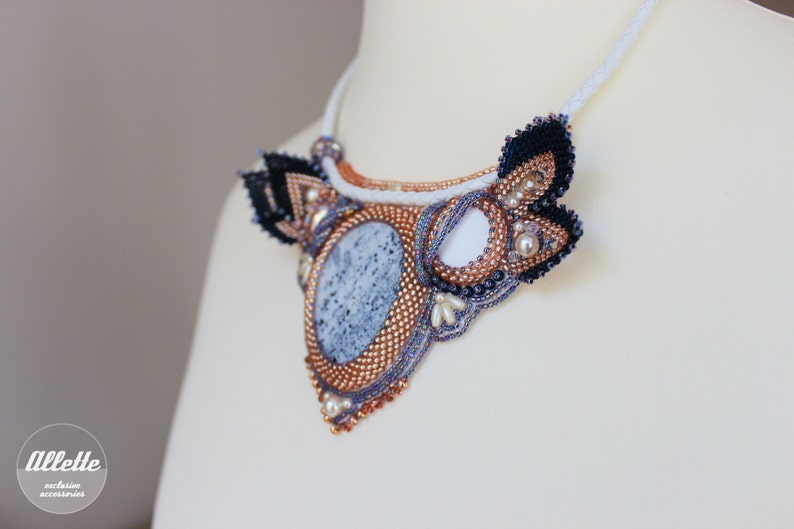 May include: A beaded necklace with a large, oval, gray and white stone pendant. The pendant is surrounded by copper-colored beads and black and white beaded wings. The necklace is on a white mannequin.