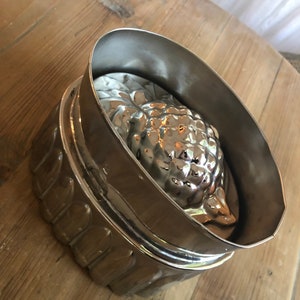 May include: A silver metal pineapple mold for making desserts. The mold is made of two pieces, a top and a bottom, and has a detailed pineapple design.