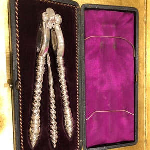 May include: A silver nutcracker and a silver nut pick in a black velvet lined box. The nutcracker has a twisted handle and a decorative top.