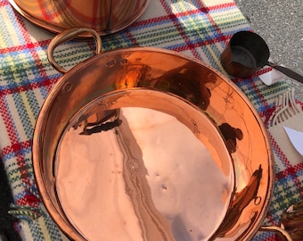 May include: A large copper serving dish with two handles. The dish is empty and has a shiny, reflective surface. The dish is sitting on a plaid blanket.