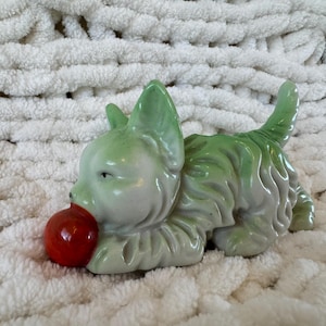 May include: A ceramic figurine of a small dog with a light green glazed finish and textured fur detail. The dog is in a playful crouching position with its tail raised and is holding a smooth, round, bright red ball in its mouth.