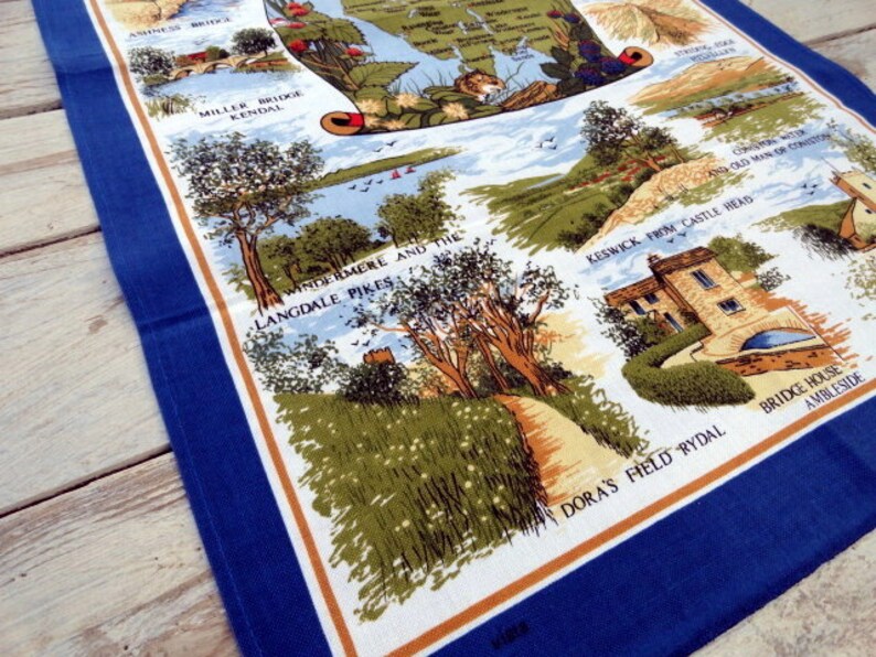 May include: A blue and white tea towel with a map of the Lake District in England. The map features various landmarks and locations, including Windermere and the Langdale Pikes, Keswick from Castle Head, and Dora's Field Rydal. The tea towel has a blue border and a brown and white pattern.