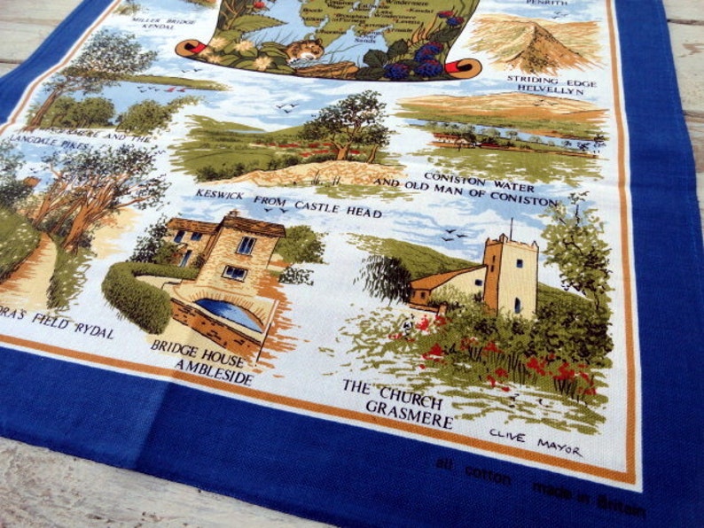 May include: A white tea towel with a blue border features a colorful illustration of the Lake District in England. The illustration includes landmarks like Helvellyn, Coniston Water, and Grasmere. The text "all cotton made in Britain" is printed in the bottom right corner.