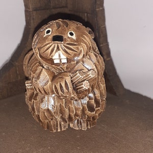 May include: A detailed, brown wooden carving of a beaver holding a bundle of sticks. The beaver has large, white teeth and small, black eyes. The carving has a textured surface and is set against a neutral background.
