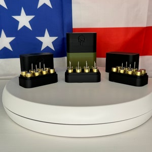 May include: Three black 3D printed boxes with gold-colored cartridges inside. The boxes are arranged in a row on a white circular surface. The box in the center has a skull logo on the front.