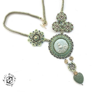 May include: A green and silver beaded necklace with a large, round pendant featuring a green and white cameo. The pendant is surrounded by smaller green and silver beads. The necklace is finished with a green and silver heart-shaped bead.