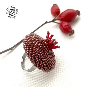 Könnte beinhalten: Handgefertigter Ring mit einem Perlendesign in Braun- und Rottönen. Der Ring hat oben eine Ansammlung roter Perlen, die einer Blume ähneln. Das verstellbare Silberband sorgt für einen modernen Touch. Der Ring liegt neben einem Zweig mit roten Beeren.