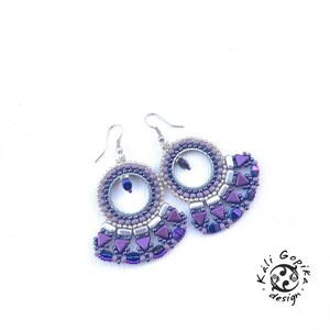 May include: Purple and silver beaded earrings with a circular design and a fan-shaped dangle. The earrings are made with small beads and have a silver hook.